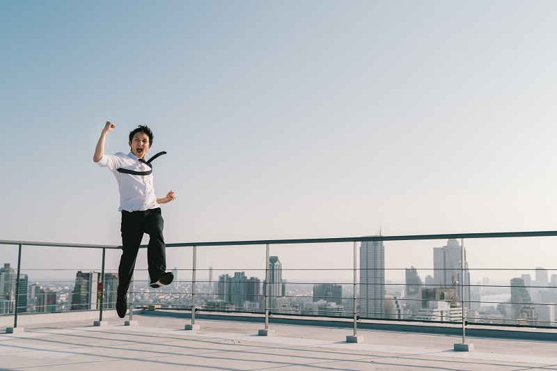 Young handsome Asian businessman jumping high, celebrate success winning pose on building rooftop. Work, job, or successful business concept. Cityscape background with copy space on sunny blue sky